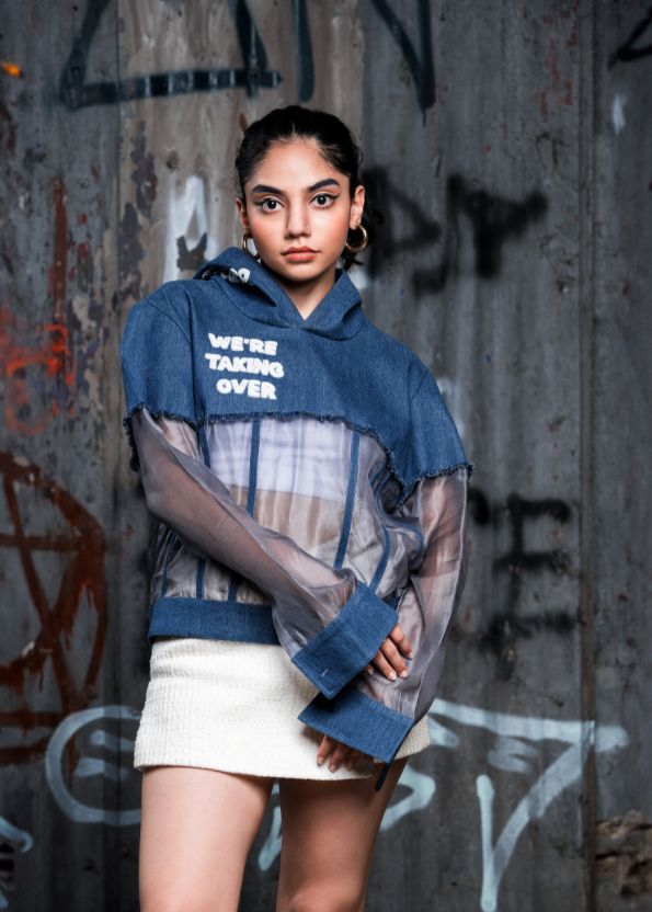 Person wearing a blue hoodie with text, sheer top, and white skirt against a graffiti-covered wall.