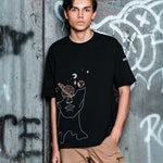 Person wearing a black t-shirt with a graphic design in front of a graffitied wall.