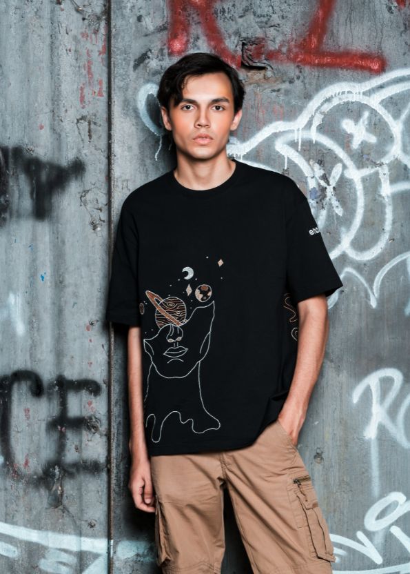 Person wearing a black t-shirt with a graphic design in front of a graffitied wall.