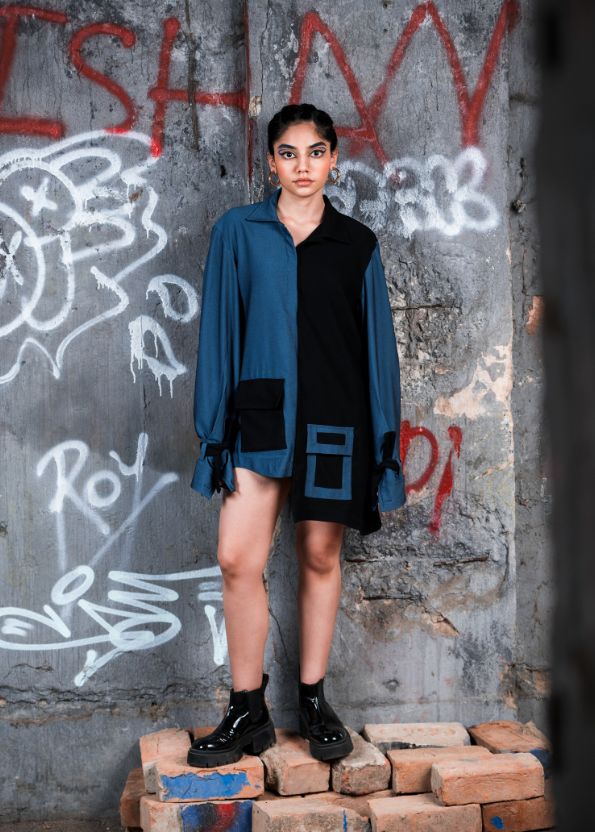 Person wearing a blue shirt over a black dress against a graffiti-covered wall.