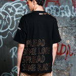 Person wearing a black t-shirt with text on the back against a graffiti-covered wall.