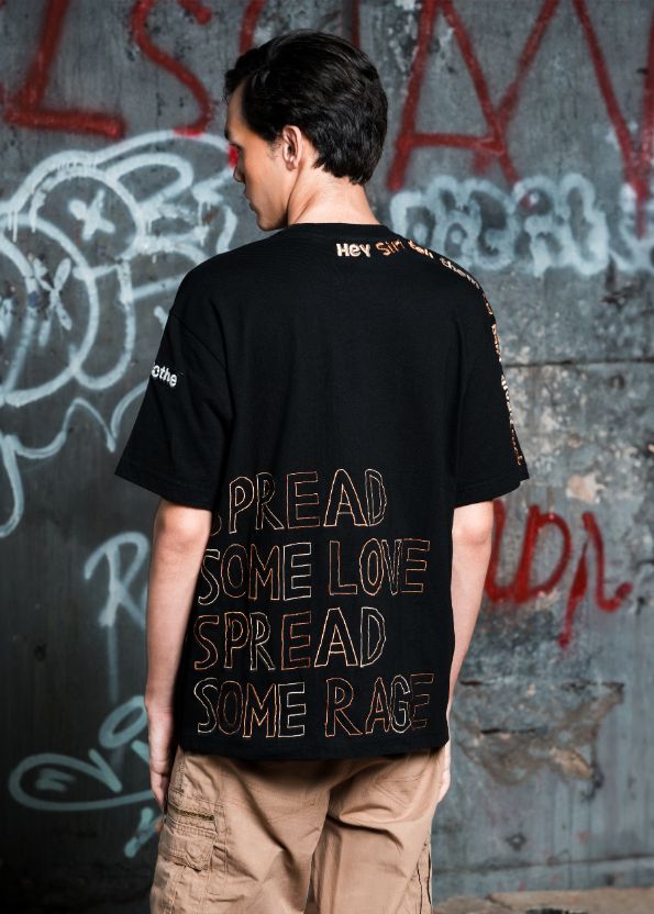 Person wearing a black t-shirt with text on the back against a graffiti-covered wall.