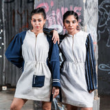 Two women wearing matching white dresses with blue accents against a graffiti-covered wall.