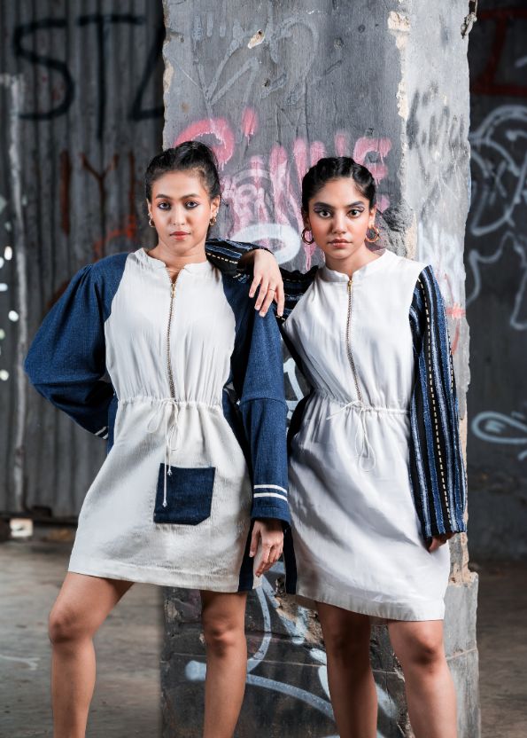 Two women wearing matching white dresses with blue accents against a graffiti-covered wall.