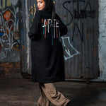 Person wearing a black coat with colorful text, standing in front of a graffiti-covered wall.