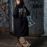 Person wearing a black coat with colorful text, standing in front of a graffiti-covered wall.