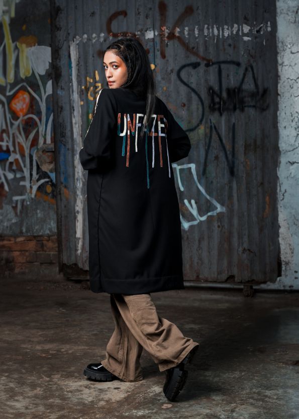 Person wearing a black coat with colorful text, standing in front of a graffiti-covered wall.
