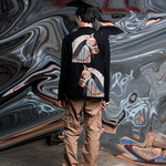Person wearing a black hoodie with colorful designs standing in front of a large abstract mural.