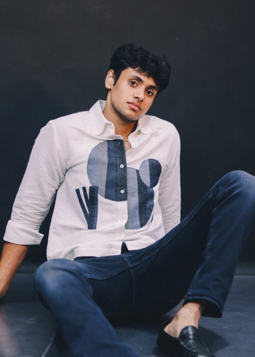 Man wearing a white shirt with blue geometric patterns and dark pants sitting against a black background