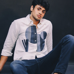Man wearing a white shirt with blue geometric patterns and dark pants sitting against a black background