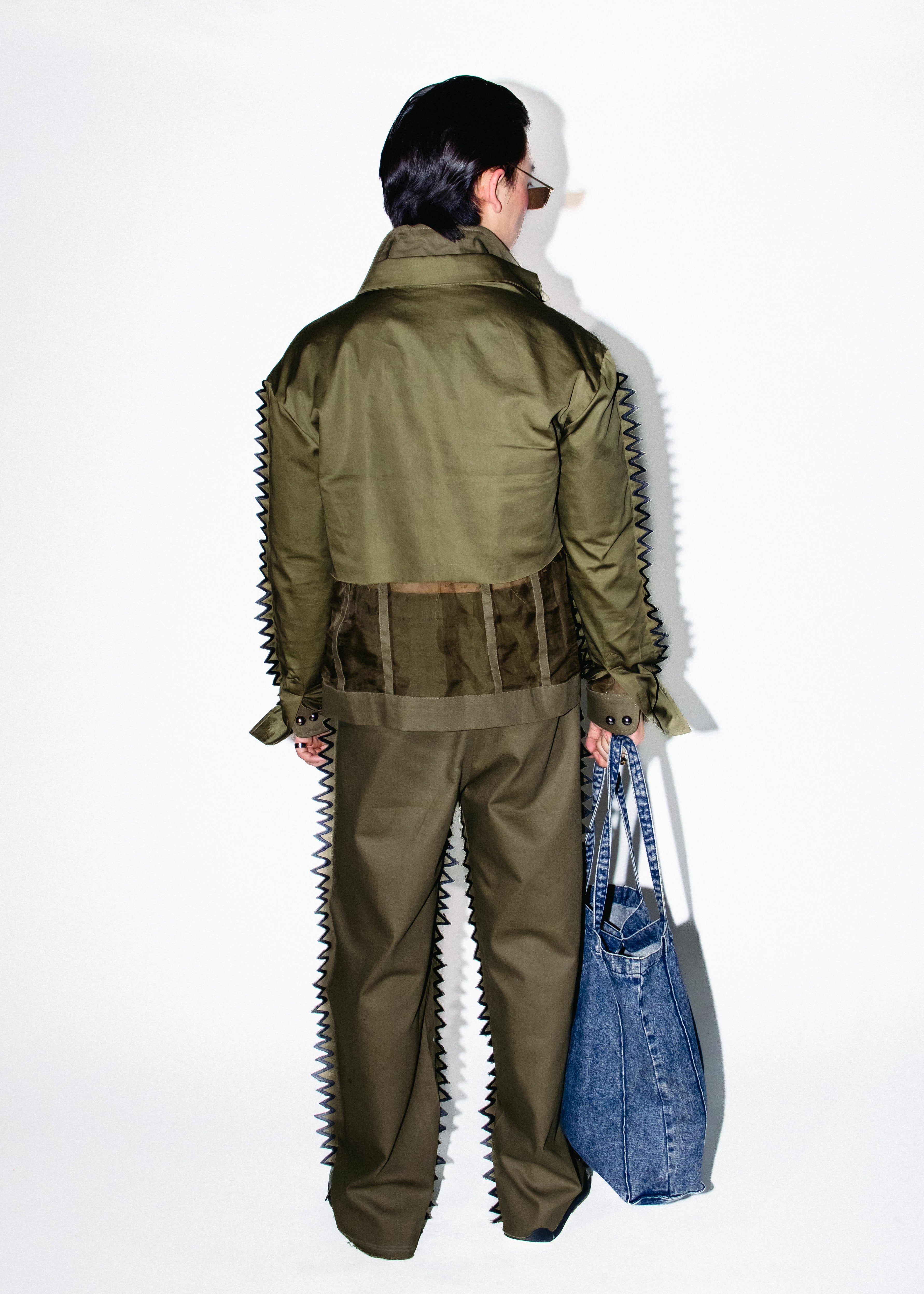 Person wearing a green military-style outfit with spiky details, holding a blue bag on a white background.