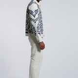 Man wearing a patterned jacket and white pants on a gray background