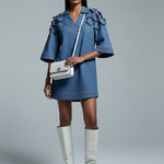 Woman wearing a blue dress and white knee-high boots holding a white handbag on a gray background