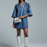 Woman wearing a blue dress and white knee-high boots holding a white handbag on a gray background
