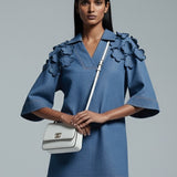 Woman wearing a blue dress with ruffled sleeves holding a white handbag on a gray background