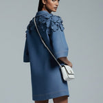 Woman wearing a blue dress with ruffled details, holding a white handbag on a gray background