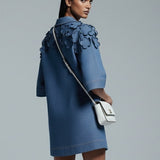 Woman wearing a blue dress with ruffled details, holding a white handbag on a gray background