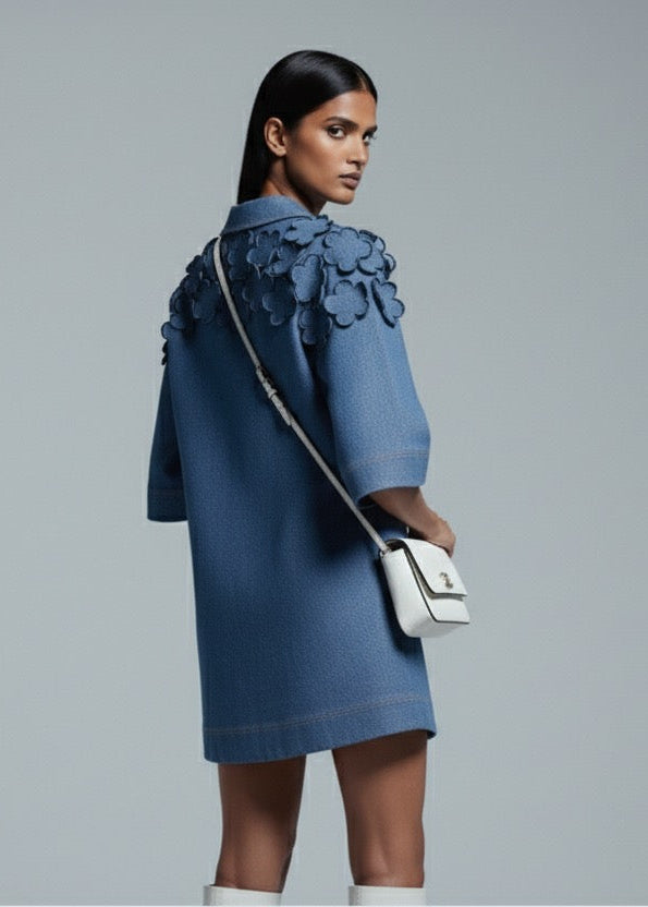 Woman wearing a blue dress with ruffled details, holding a white handbag on a gray background