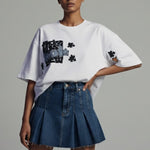 Woman wearing a white oversized t-shirt with graphic designs and a blue denim skirt on a gray background