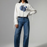 Woman wearing a white blouse with a blue design and blue jeans on a gray background