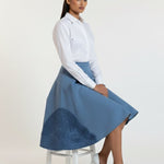 Woman wearing a white blouse and blue skirt sitting on a stool against a white background