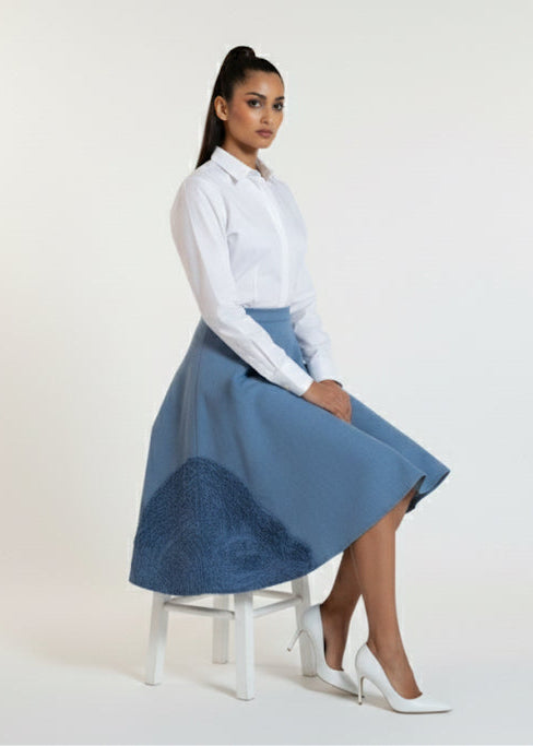 Woman wearing a white blouse and blue skirt sitting on a stool against a white background