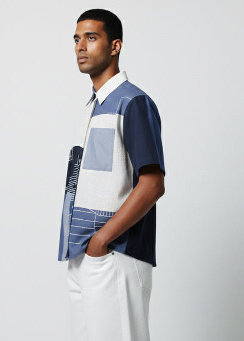 Man wearing a patterned shirt and white pants against a plain background