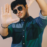 Man wearing sunglasses and a two-tone shirt with arms raised against a beige background