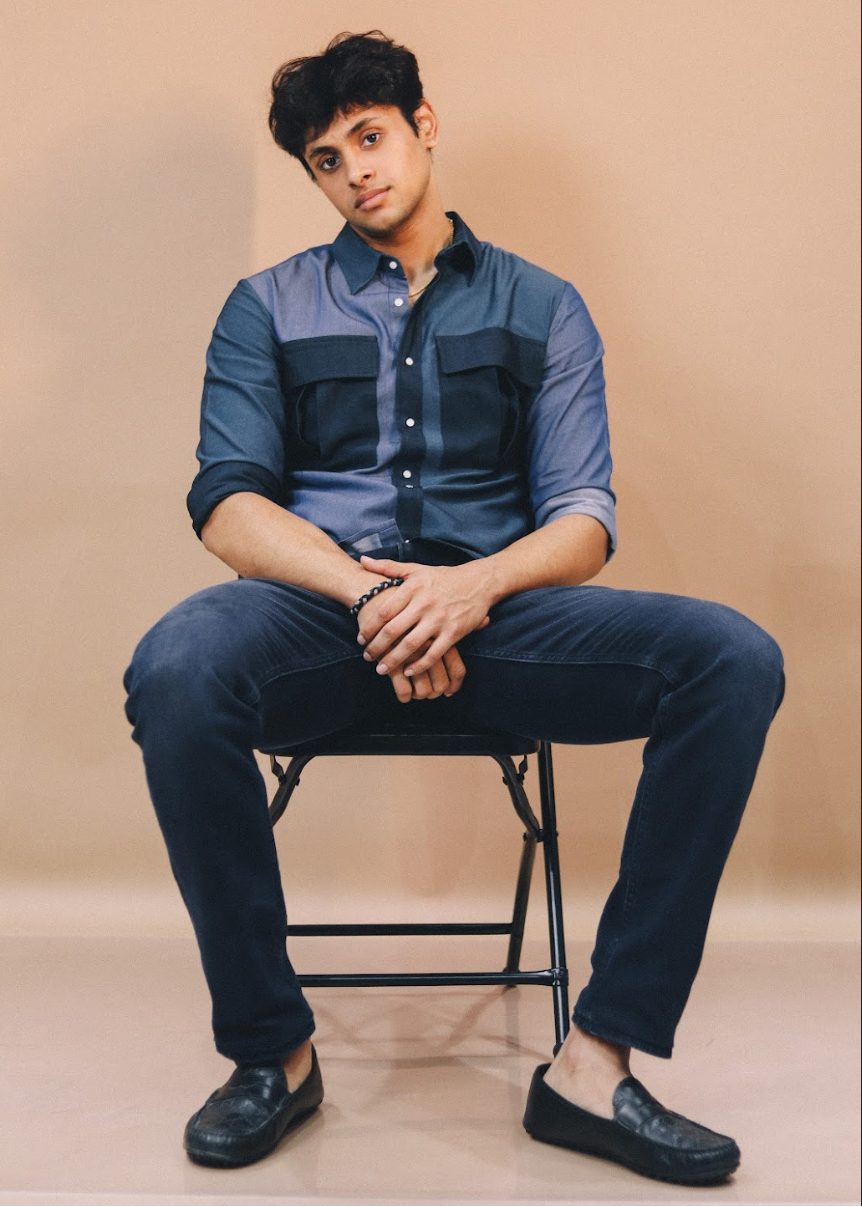 Man sitting on a chair wearing a blue shirt and dark pants against a beige background