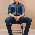 Man sitting on a chair wearing a blue shirt and dark pants against a beige background